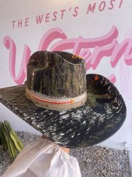 Camo cowboy hat with neon orange stitching and frayed beige band, held against a pink Western mural and desert cactus backdrop.