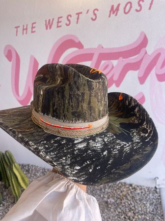 Camo cowboy hat with neon orange stitching and frayed beige band, held against a pink Western mural and desert cactus backdrop.