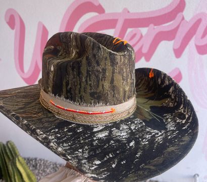 Camo cowboy hat with neon orange stitching and frayed beige band, held against a pink Western mural and desert cactus backdrop.