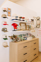 Bright boutique hat display with stacked baseball caps and a red trucker hat on white shelves, bracelets and jewelry in frames, enamel patches and pins in clear trays atop a light-wood dresser, vintage racing sign and wall mirror accents.
