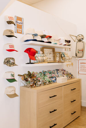 Bright boutique hat display with stacked baseball caps and a red trucker hat on white shelves, bracelets and jewelry in frames, enamel patches and pins in clear trays atop a light-wood dresser, vintage racing sign and wall mirror accents.