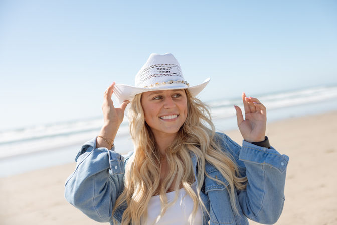 Smiling woman with long blonde hair wearing a white straw cowboy hat and denim jacket on a sunny sandy beach by the ocean