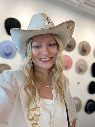 Smiling woman wearing a white embroidered cowboy hat and gold-letter sash in a western hat boutique, colorful cowboy hats displayed on the wall behind her