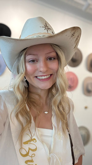 Smiling woman wearing a white embroidered cowboy hat and gold-letter sash in a western hat boutique, colorful cowboy hats displayed on the wall behind her