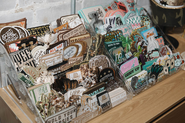 Close-up of acrylic display racks on a wooden counter against a white brick wall holding colorful embroidered patches and novelty pins — Western, retro and playful designs like boots, horses and fishing motifs.