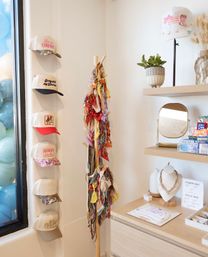Light-filled boutique accessory display with wall-mounted trucker hats, a wooden stand of colorful bandanas, small jewelry display, round mirror and potted plant