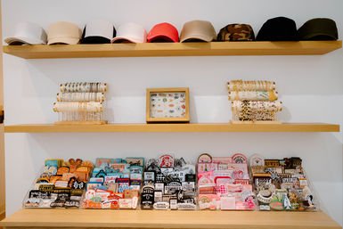 Boutique wooden shelves showing a row of baseball caps on the top shelf, bracelet stands and a framed pin collection on the middle shelf, and colorful enamel pins and patches neatly organized in trays on the bottom — trendy retail accessory display.