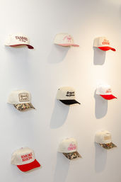 Nine white baseball caps with colorful brims and embroidered slogans arranged in a 3x3 grid on a white wall display — boutique-style hat display with red, pink, black and camouflage brims.