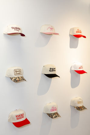 Nine white baseball caps with colorful brims and embroidered slogans arranged in a 3x3 grid on a white wall display — boutique-style hat display with red, pink, black and camouflage brims.