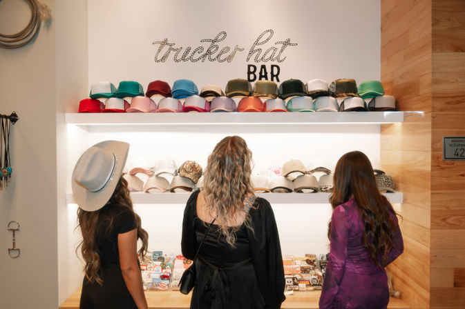 Three women seen from behind browsing a colorful trucker hat bar display in a bright boutique hat shop, rows of multicolored caps on white shelves with warm wood accents.