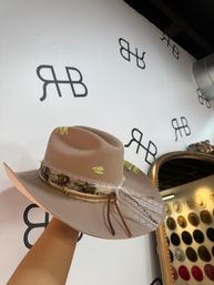 Tan felt boho fedora with feather, lace and braided leather band held up in a hat boutique against a white wall with repeating logo and a framed mirror showing a hat display.
