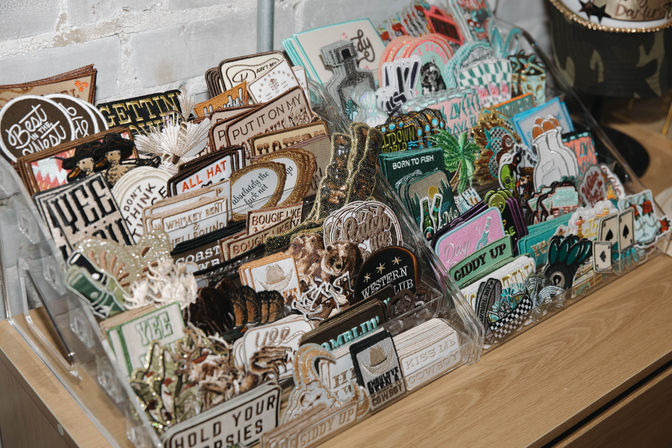 Clear acrylic display rack of colorful embroidered patches with cowboy, animal, and novelty designs on a wooden counter by a white brick wall.