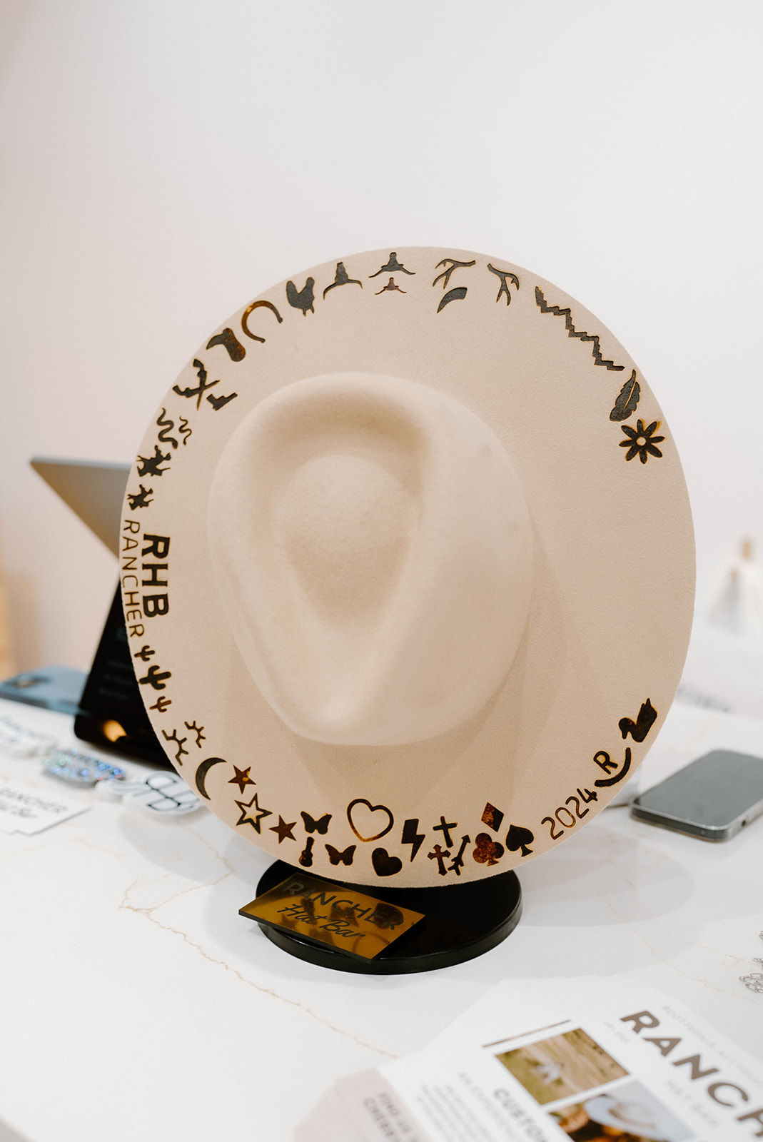 Beige wide-brim felt cowboy hat on a tabletop stand, decorated with burned playful icons (hearts, stars, cacti, animals) and a '2024' mark along the brim, retail display.