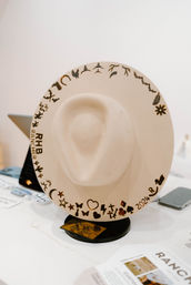 Beige wide-brim felt cowboy hat on a tabletop stand, decorated with burned playful icons (hearts, stars, cacti, animals) and a '2024' mark along the brim, retail display.