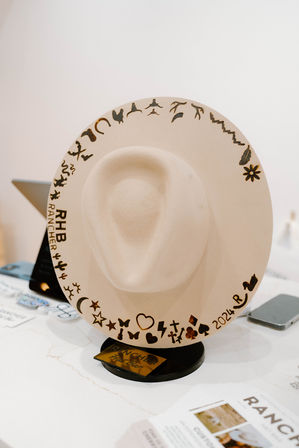 Beige wide-brim felt cowboy hat on a tabletop stand, decorated with burned playful icons (hearts, stars, cacti, animals) and a '2024' mark along the brim, retail display.