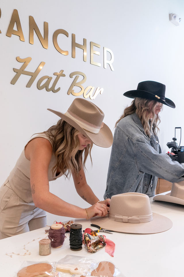 Two women in a boutique hat shop customizing wide-brim felt cowboy hats at a white counter with ribbons, spools and hatmaking tools beneath gold wall lettering.
