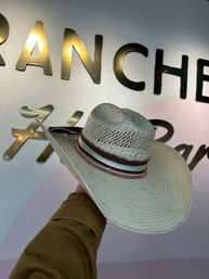 Straw cowboy hat with wide brim and pink-and-brown decorative band held up against a white wall featuring gold metallic letters in a retail shop display.