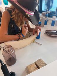 Person wearing a cowboy-style wide-brimmed hat hand-tooling and decorating a felt hat brim at a DIY hat-making craft studio counter with torches, tools, a mason jar, and wooden blocks.