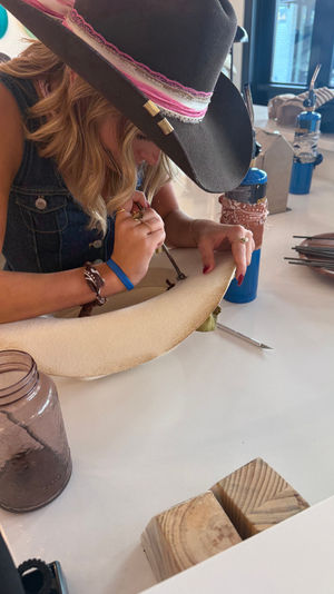 Person wearing a cowboy-style wide-brimmed hat hand-tooling and decorating a felt hat brim at a DIY hat-making craft studio counter with torches, tools, a mason jar, and wooden blocks.