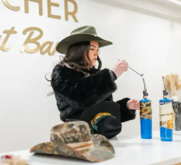 Woman in a green wide-brim hat and black faux-fur coat using a handheld torch at an artisan hat workshop, cowboy hats on the counter and blue propane torches nearby.