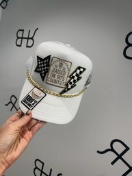 White mesh-back trucker hat held by a hand, embroidered patches including "Raised on 90's Country", black-and-white checkerboard star and lightning bolt, gold chain trim and curved brim on a neutral backdrop