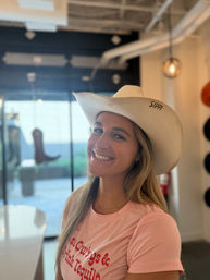 Smiling woman in a pink t-shirt wearing a white cowboy hat inside a bright hat boutique, with a boot display and hanging pendant lights in the blurred background.