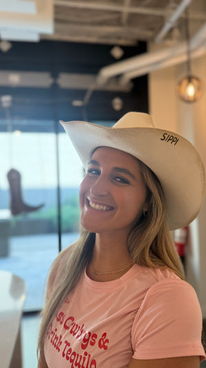 Smiling woman in a pink t-shirt wearing a white cowboy hat inside a bright hat boutique, with a boot display and hanging pendant lights in the blurred background.