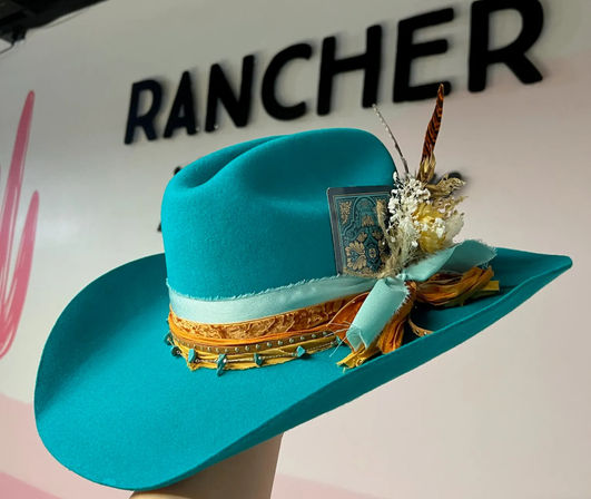 Vibrant turquoise felt cowboy hat with wide brim, layered orange and pale-blue ribbons, turquoise studs, dried flowers and a pheasant feather accent, photographed against a pale wall with bold black lettering.