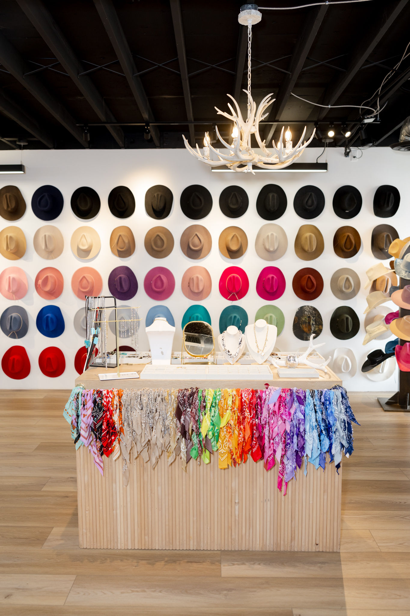 Boutique interior with a colorful wall of hats, a wooden display table draped with rainbow bandanas and jewelry, and an antler chandelier overhead.