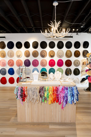 Boutique interior with a colorful wall of hats, a wooden display table draped with rainbow bandanas and jewelry, and an antler chandelier overhead.