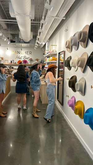 Group of shoppers trying on western-style cowboy hats in a modern hat boutique with a colorful wall display of felt hats, mirrors, wooden counters and polished concrete floors