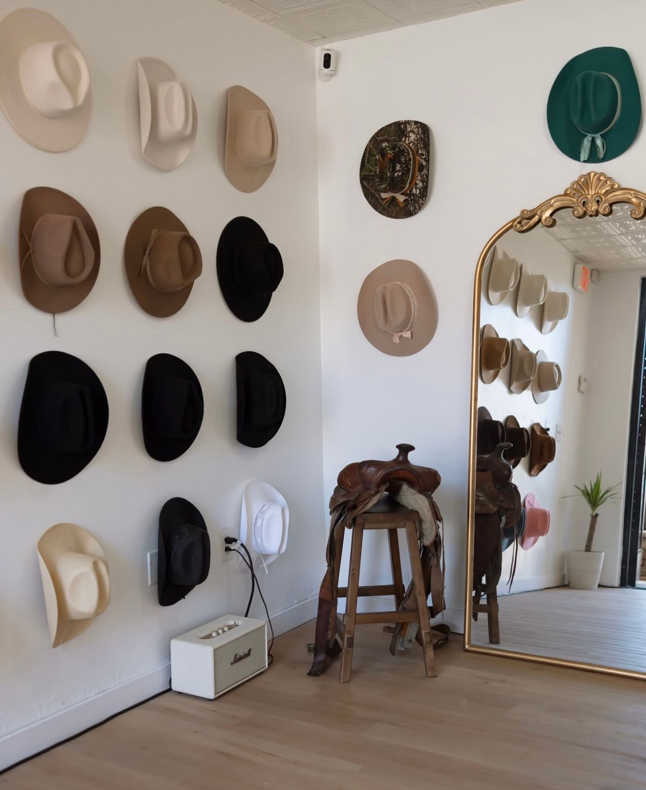 Western hat wall display with neutral, black, and teal cowboy hats beside a full-length gold mirror, wooden stool holding a leather saddle, white speaker on the floor, and a small potted plant in a bright boutique interior.