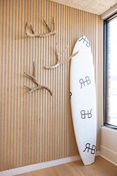 White surfboard leaning against a vertical wood-slat wall with three decorative antlers beside a sunlit window, minimalist coastal interior decor.
