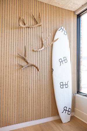 White surfboard leaning against a vertical wood-slat wall with three decorative antlers beside a sunlit window, minimalist coastal interior decor.