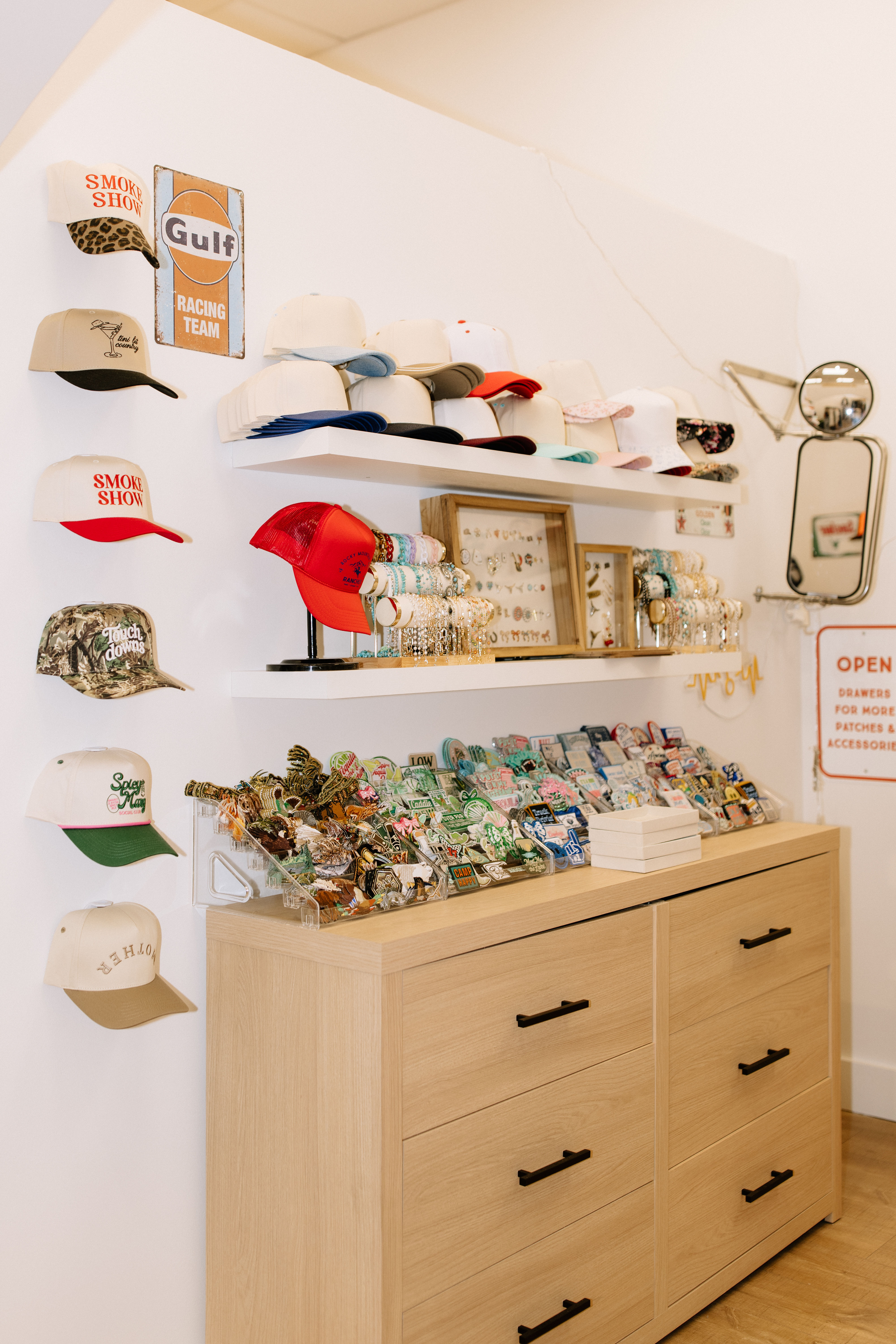 Bright boutique wall display of stacked baseball caps, a red trucker hat on a stand, bracelets and framed earrings, and trays of enamel pins and patches atop a light oak dresser.