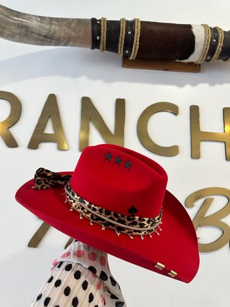 Red cowboy hat with leopard-print band, dangling star charms and three black stars on the crown, held against a white wall with gold ranch-style letters and a mounted horn