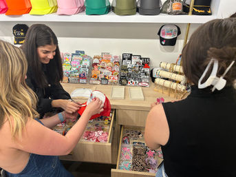 Three women shopping a boutique accessory display — one holding a red trucker hat while others browse colorful caps, enamel patches, pins and bracelet racks.