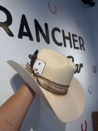 Beige felt cowboy hat with feather and layered ribbon band, a 3 of diamonds playing card peeking from the band, held against a Western-themed wall decorated with horseshoes.