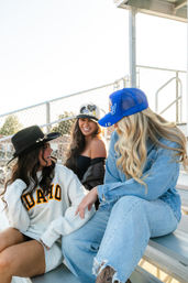 Three friends laughing on metal stadium bleachers at a sunny sports field — one wearing an 'IDAHO' sweatshirt and black hat, one in denim with a blue trucker cap, and one in a strapless top with a patch hat.