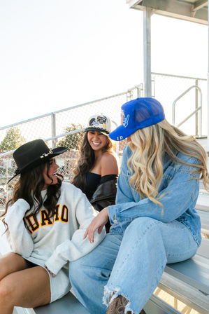 Three friends laughing on metal stadium bleachers at a sunny sports field — one wearing an 'IDAHO' sweatshirt and black hat, one in denim with a blue trucker cap, and one in a strapless top with a patch hat.
