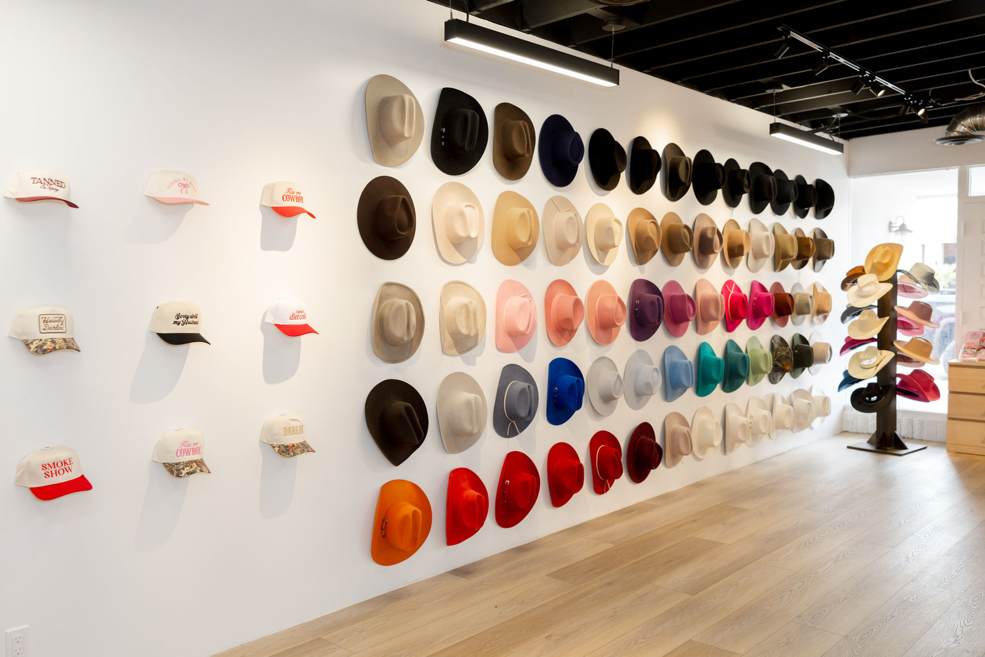 Bright hat shop interior with colorful cowboy and wide-brim hats arranged in neat rows on a white gallery wall, baseball caps on the left and a rotating rack by the window.