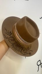Brown boho felt fedora with wide brim, black hand-drawn floral design on the brim, braided leather band and lace trim, photographed against a white background.