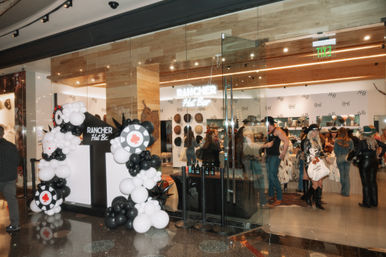 Indoor shopping mall hat shop pop-up with glass storefront and wood accents, black-and-white balloon arch with casino-chip and playing-card motifs, shoppers browsing and trying on western-style hats.