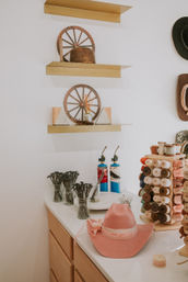 Pink cowboy hat on a white work counter surrounded by jars of hat pins, stacked spools of ribbon, two blue torch tools and decorative wooden wagon-wheel shelves in a hat-making workshop