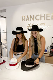 Two women wearing black cowgirl hats customizing a white and a black cowboy hat on a bright marble counter in a western-style hat bar
