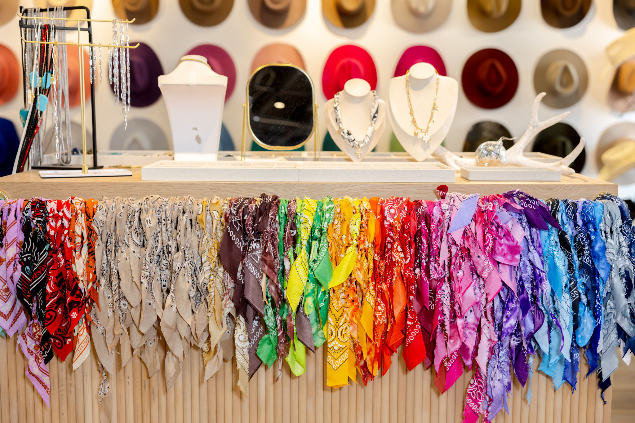 Boutique accessory display with a rainbow of colorful bandanas draped along a wooden counter, jewelry busts and mirror on the tabletop, and a wall of hats in the background.