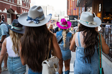 Four women in denim outfits wearing decorative cowboy hats — one bright pink — walking down a busy downtown street, braided hair and city buildings in the background.