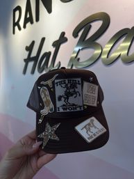 Hand holding brown trucker cap decorated with western patches — glitter star, cowboy boot, longhorn, bucking-bronco patch with bold lettering, "YEE HAW" rectangle and a dog patch — in front of a pink wall with metallic script signage.