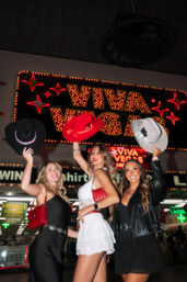 Three women posing under a glowing 'Viva Vegas' neon marquee at night, lifting black, red, and gray cowboy hats and smiling on a lively Las Vegas nightlife street, wearing stylish outfits and red purses.