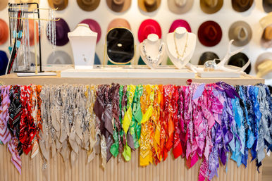 Boutique accessory display with a rainbow of colorful bandanas draped along a wooden counter, jewelry busts and mirror on the tabletop, and a wall of hats in the background.